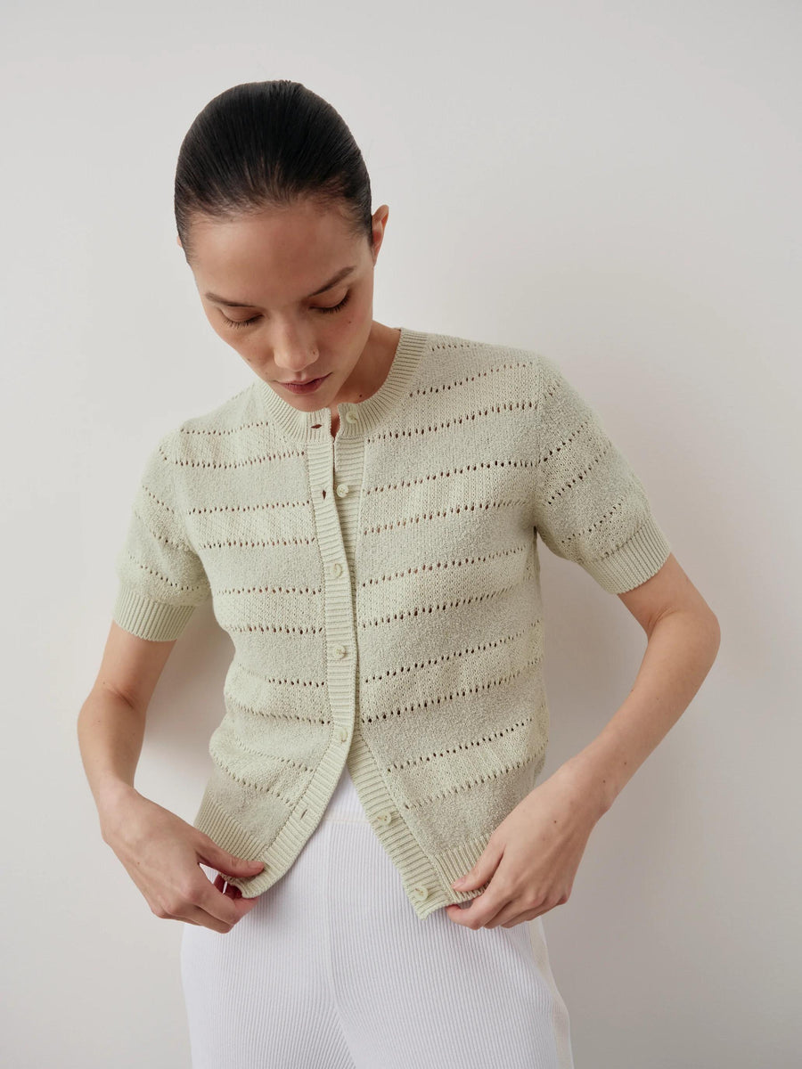 Model wearing Emello's Bouclé Short Sleeve Cardigan in pale green with delicate pointelle stripes, paired with white trousers.