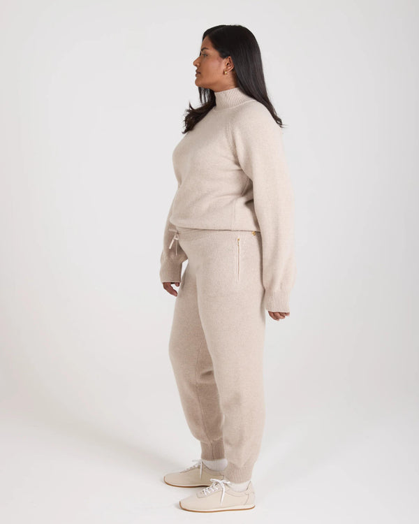 Elegant woman in beige cashmere blend knit trousers with ribbed cuffs and gold accents, standing in side profile.