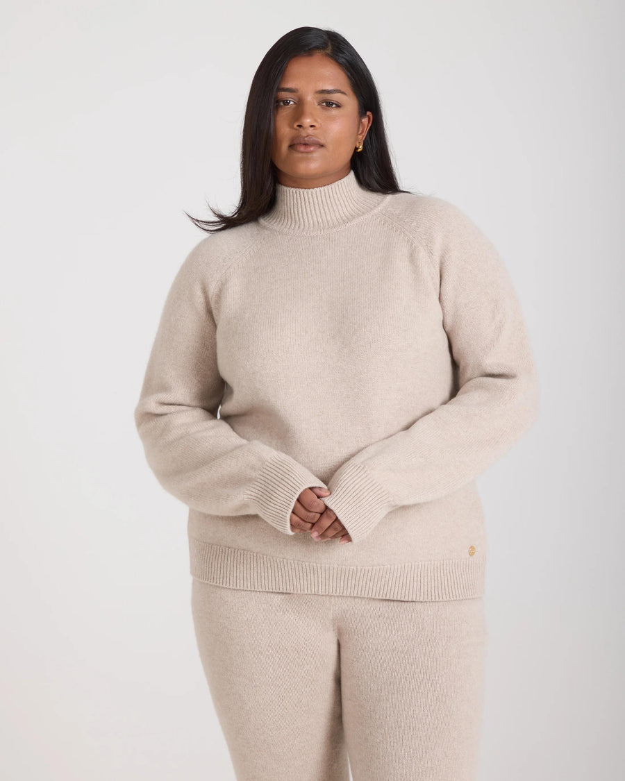 Kasima Cashmere Blend Knit Sweater in oatmeal beige, featuring a relaxed fit on a model with folded over rib funnel neck and raglan sleeves.