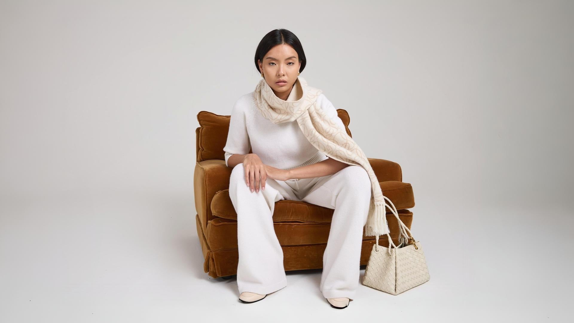 Model in soft ivory knit set with tonal scarf and woven bag seated on velvet chair