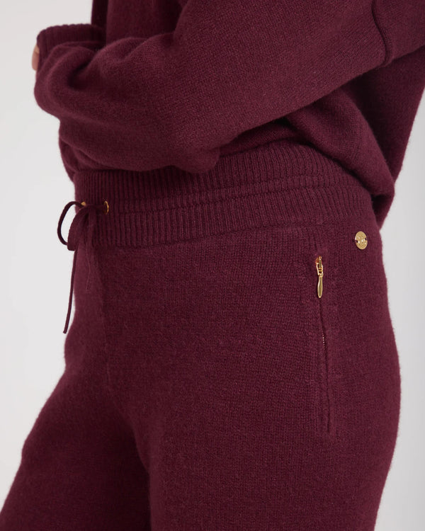 Close-up of Kasima cashmere blend knit trousers in fig burgundy with gold-finish disc and concealed zips.