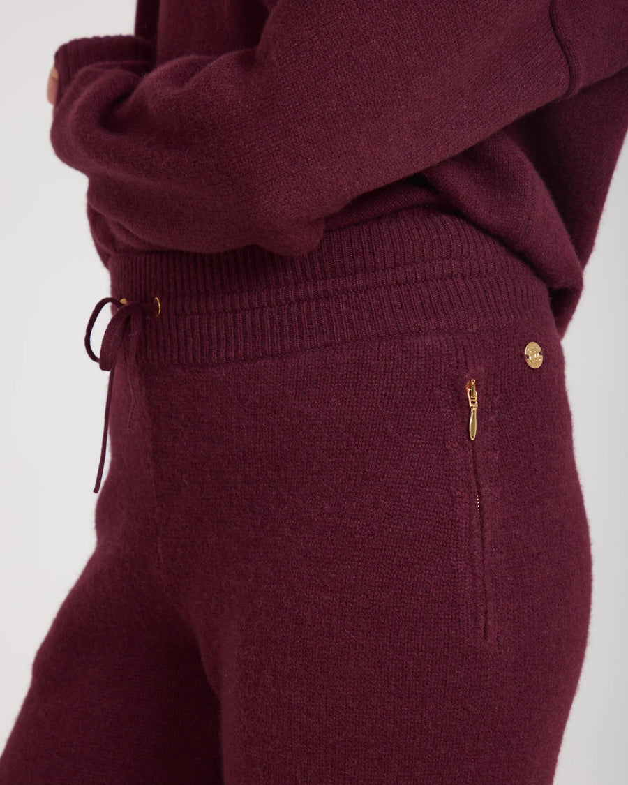 Close-up of Kasima cashmere blend knit trousers in fig burgundy with gold-finish disc and concealed zips.