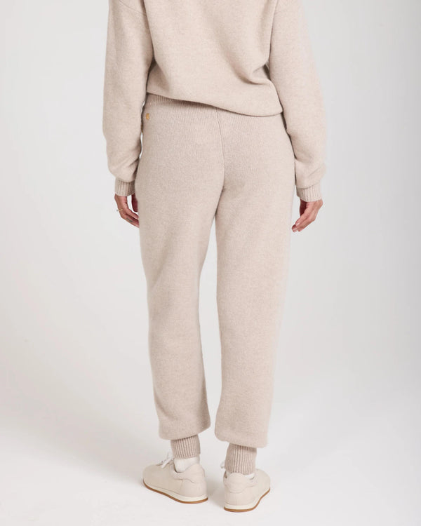 Back view of beige Kasima cashmere blend knit trousers by Emello, showcasing ribbed cuffs and gold disc embellishment.