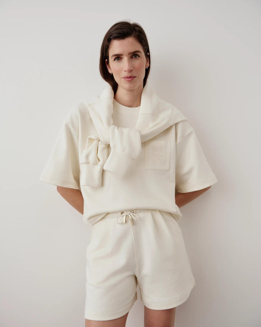 Model wearing Emello embroidered pocket shorts in cream with a matching sweatshirt draped over the shoulders.