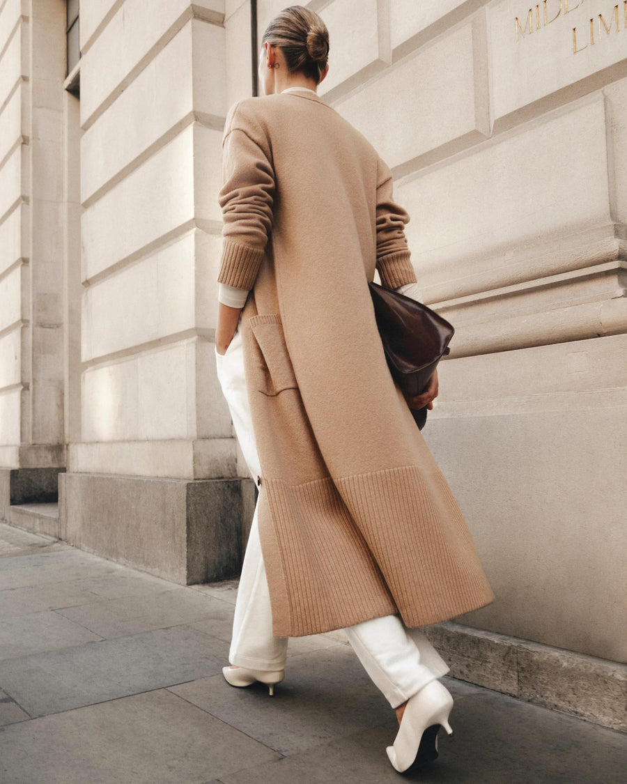 Model wearing a long brown merino wool cardigan by Emello, walking alongside a building with elegant architecture.