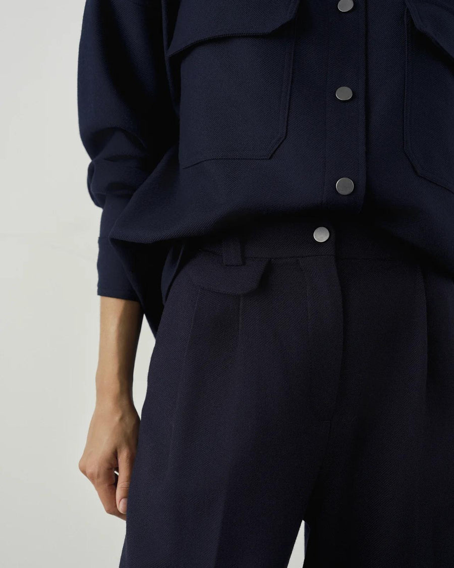 Pleated wool twill trousers in deep navy with front pleats and brushed-metal fastening, styled with coordinating overshirt.