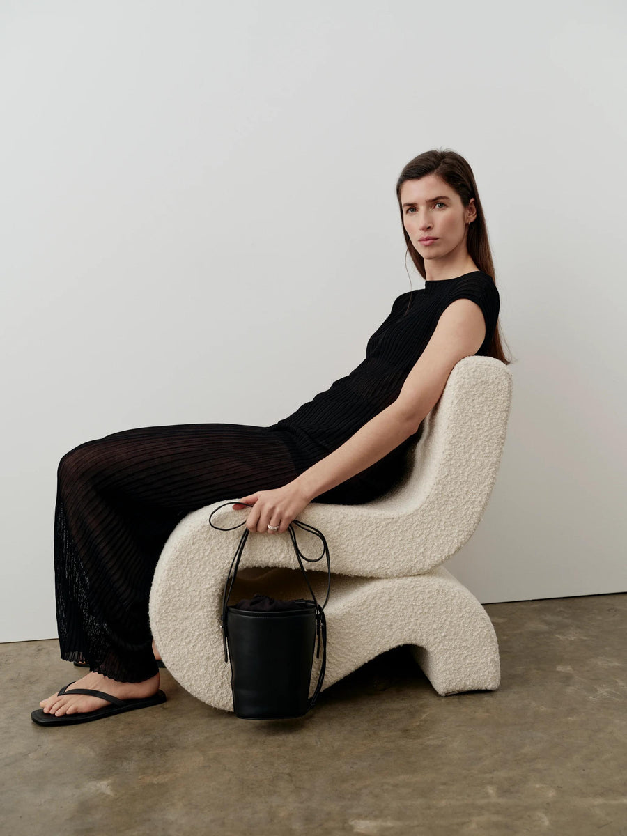 Model wearing black plissé knit trousers seated on a modern boucle chair, showcasing the fluid and light-catching design.