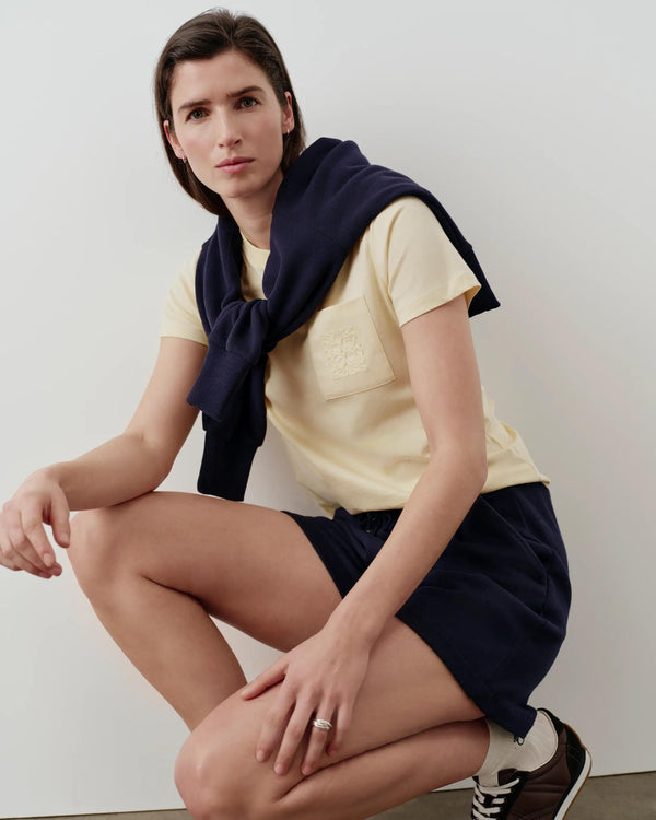 A model wearing a butter-yellow Supima cotton pocket tee with a navy sweater draped over shoulders, showcasing Emello's signature elegance.
