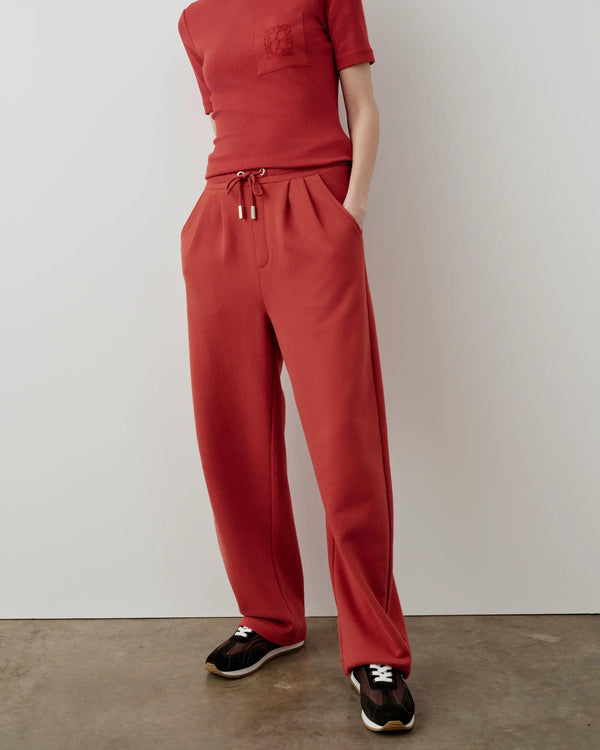 A model wearing relaxed straight leg trousers in muted rust red, featuring a drawcord waistband and side pockets.