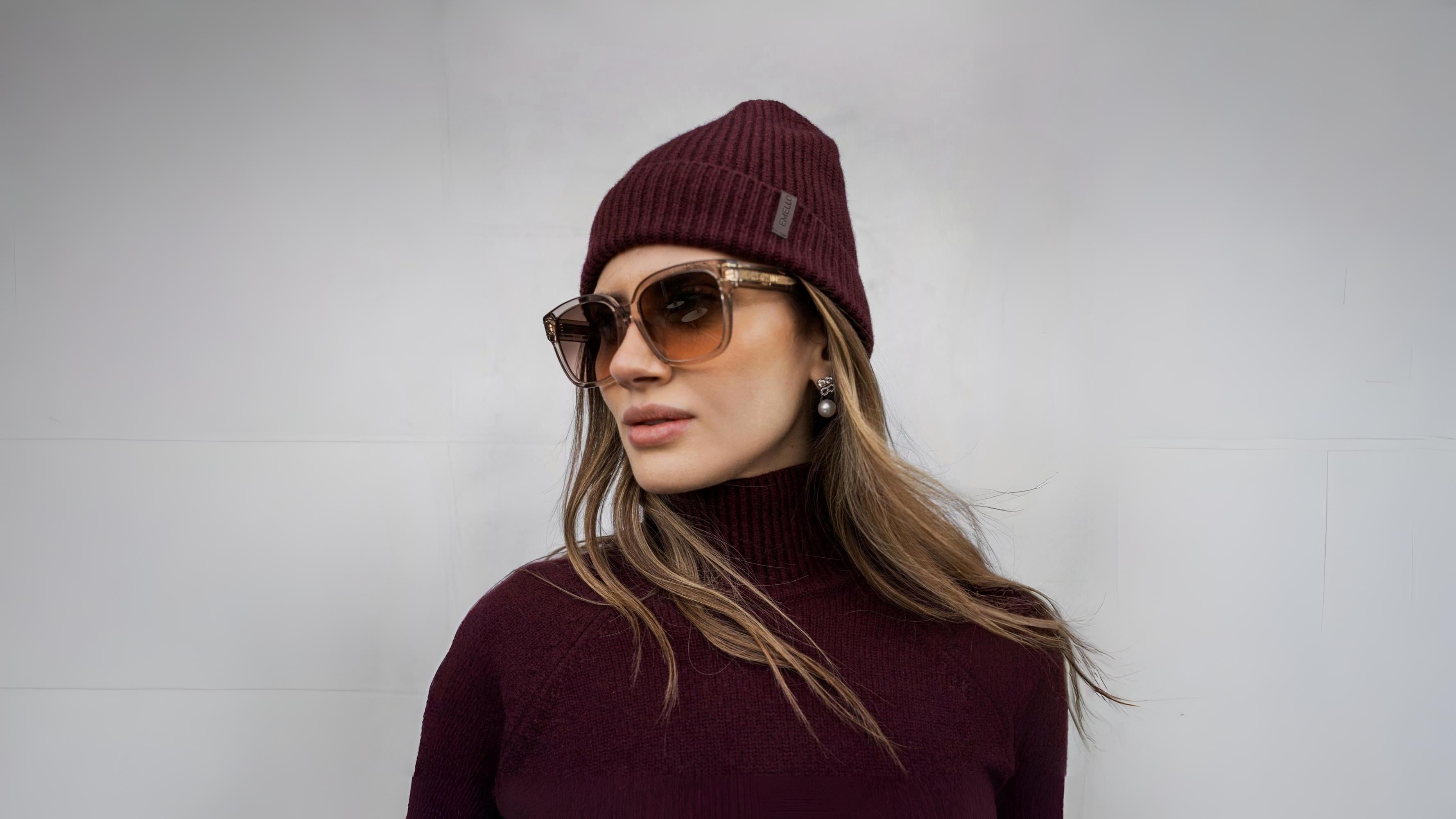 Woman wearing a ribbed burgundy cashmere beanie and matching knit, styled with oversized sunglasses and pearl earrings.