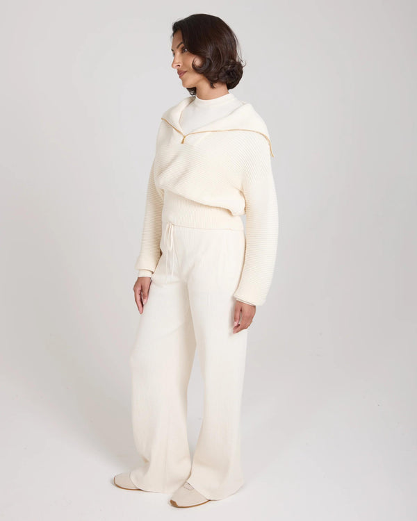 Elegant woman wearing a cream ribbed half-zip jumper, standing with relaxed poise, set against a soft, neutral background.