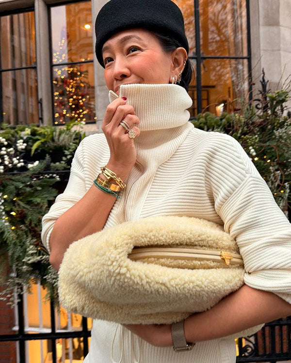 Woman in ribbed ivory knit holding a shearling clutch, wearing layered gold jewelry