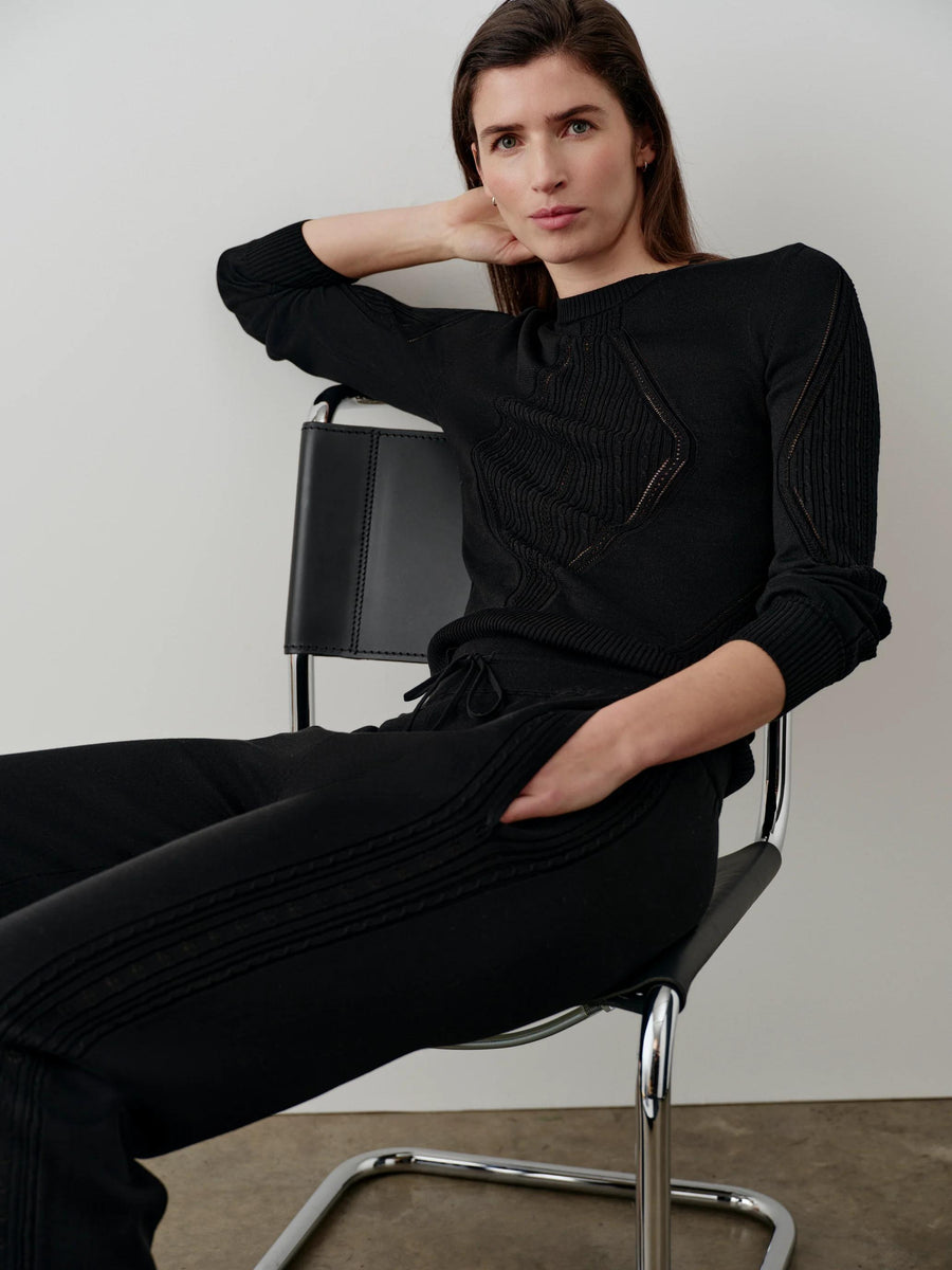 Model wearing a black silk blend pointelle sweater with geometric designs and blouson sleeves, seated in a modern chair.