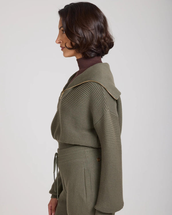 Side view of a woman wearing the Sorella Ribbed Half Zip Jumper in Olive, showcasing its relaxed fit and gold zip details.