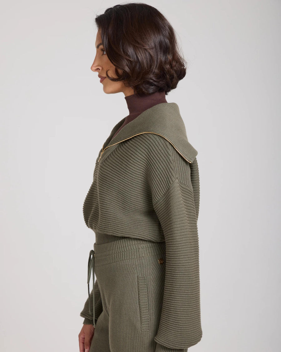 Side view of a woman wearing the Sorella Ribbed Half Zip Jumper in Olive, showcasing its relaxed fit and gold zip details.