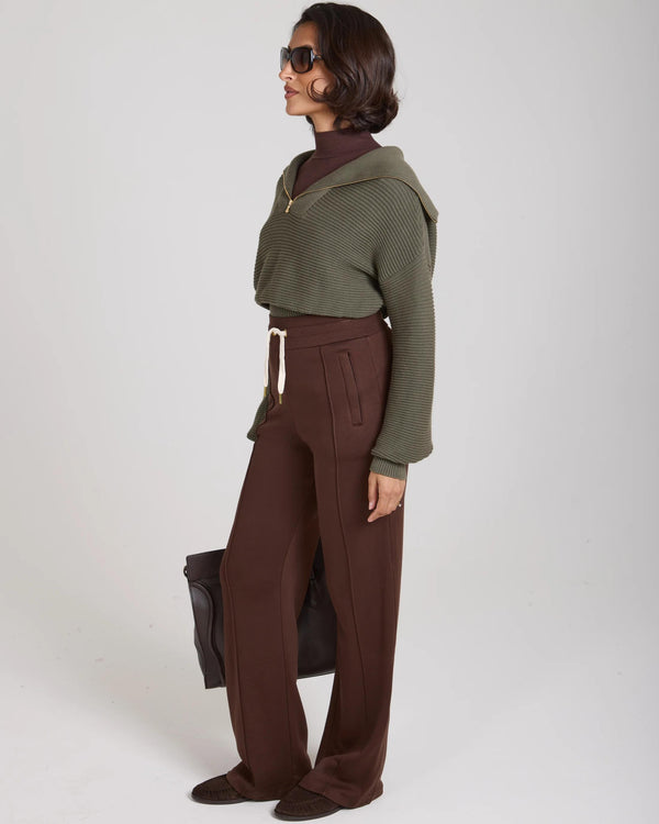 Elegant woman wearing Emello's olive Sorella ribbed half zip jumper, layered over a chocolate brown turtleneck, styled with brown wide-leg trousers, embodying refined ease and tactile elegance.