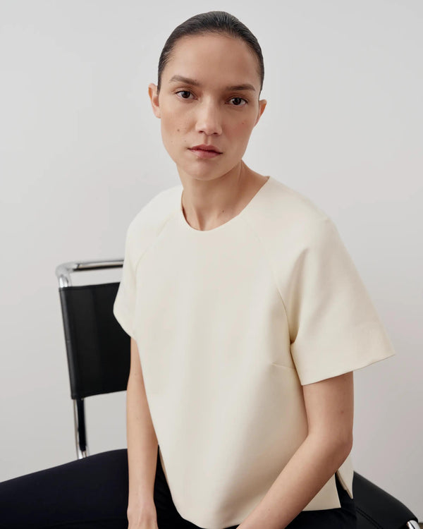 Woman wearing a cream structured short sleeve top with a sculpted fit, seated on a modern chair.
