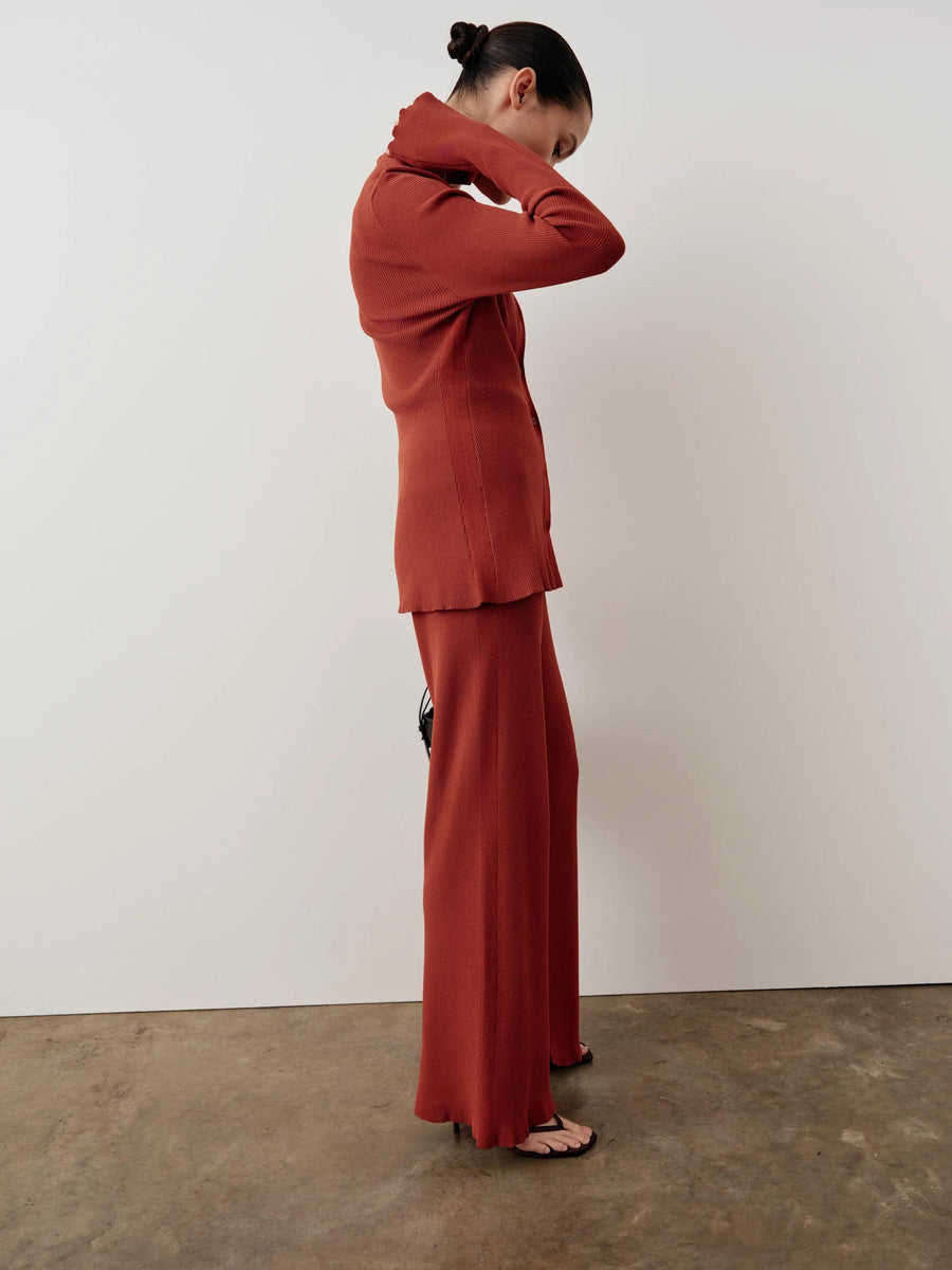 Side profile of a model wearing Emello's tonal ribbed knit trousers in warm rust red, illustrating fluid movement.